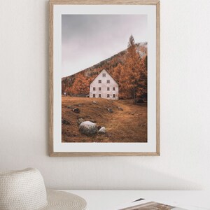 May include: A framed photograph of a white house with multiple windows, nestled in a hillside covered in autumn leaves. The house is surrounded by trees and rocks, with a cloudy sky in the background.