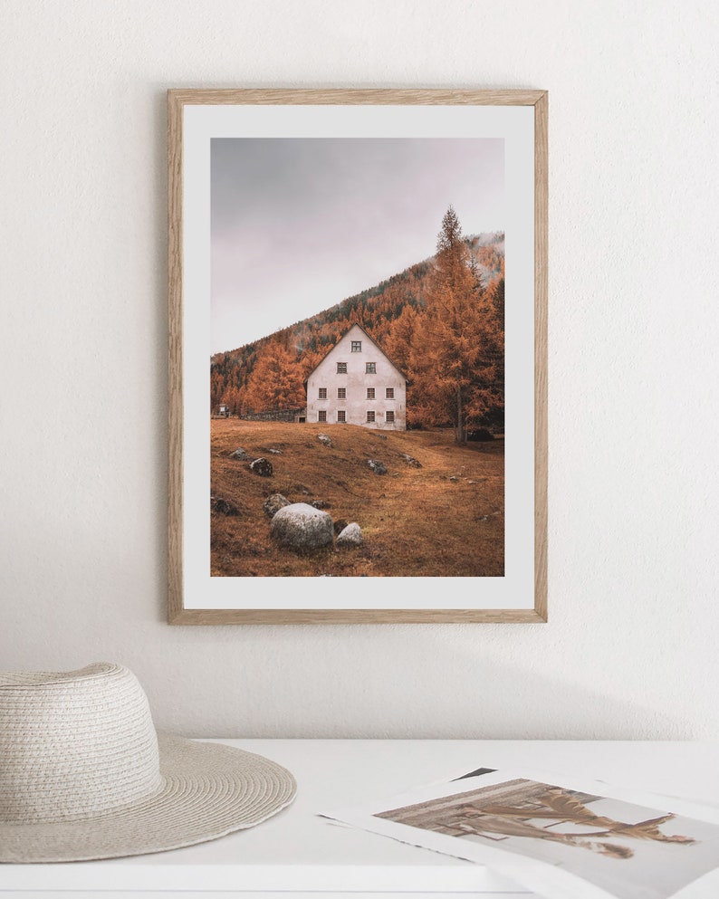 May include: A framed photograph of a white house with multiple windows, nestled in a hillside covered in autumn leaves. The house is surrounded by trees and rocks, with a cloudy sky in the background.