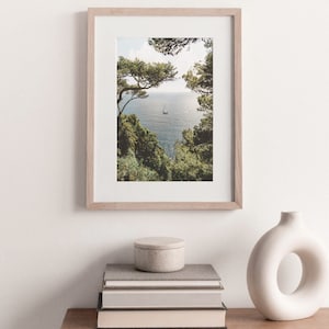 K&ouml;nnte beinhalten: Ein gerahmtes Foto eines Segelboots auf dem Wasser, das durch einen Rahmen aus B&auml;umen zu sehen ist. Das Foto h&auml;ngt an einer wei&szlig;en Wand &uuml;ber einem Stapel B&uuml;cher und einer wei&szlig;en Vase.