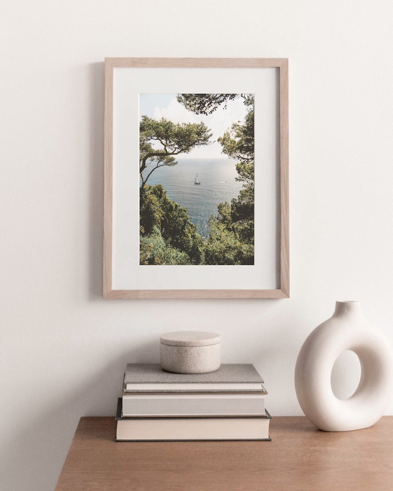 K&ouml;nnte beinhalten: Ein gerahmtes Foto eines Segelboots auf dem Wasser, das durch einen Rahmen aus B&auml;umen zu sehen ist. Das Foto h&auml;ngt an einer wei&szlig;en Wand &uuml;ber einem Stapel B&uuml;cher und einer wei&szlig;en Vase.