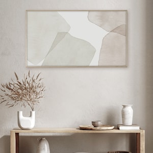 Op de afbeelding: Een ingelijst abstract aquarel schilderij met gedempte groene en bruine vormen op een witte achtergrond. Het schilderij hangt aan een witte muur boven een houten plank met decoratieve items.
