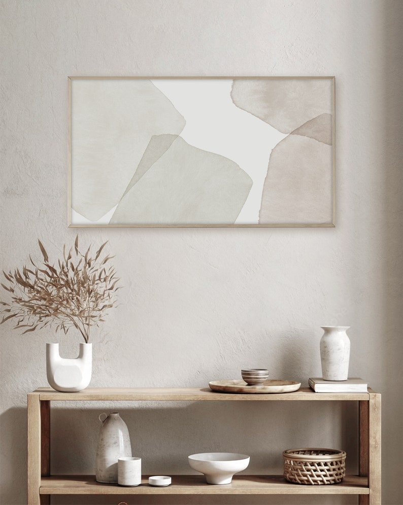 Op de afbeelding: Een ingelijst abstract aquarel schilderij met gedempte groene en bruine vormen op een witte achtergrond. Het schilderij hangt aan een witte muur boven een houten plank met decoratieve items.