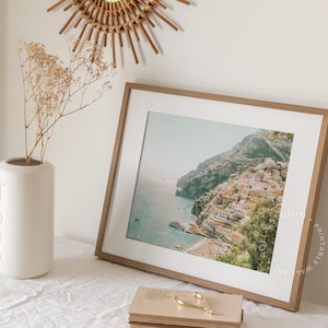 May include: A framed print of a coastal town with colorful buildings on a hillside overlooking the ocean. The print is in a light wood frame and is hanging on a white wall.