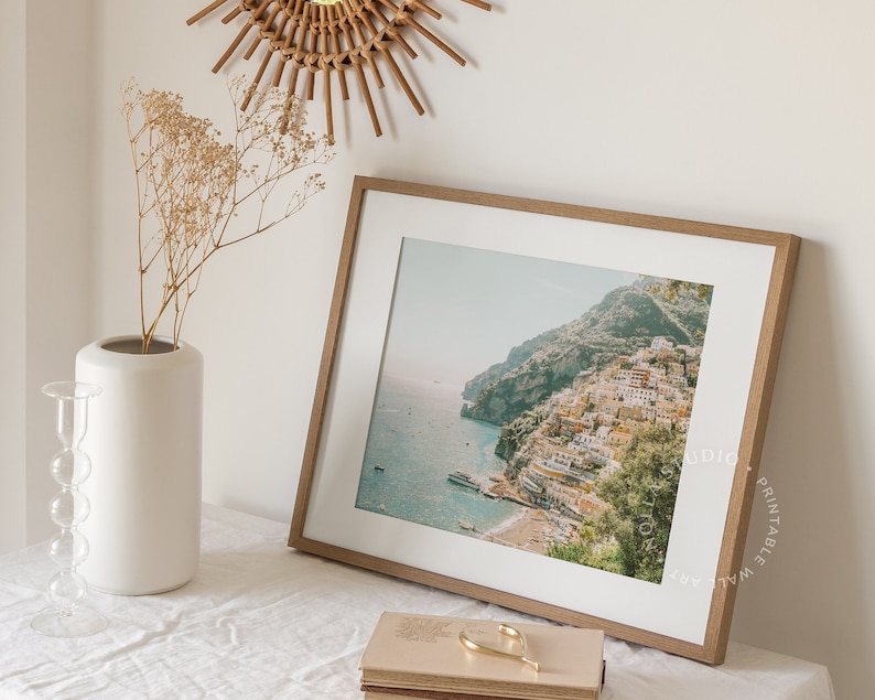 May include: A framed print of a coastal town with colorful buildings on a hillside overlooking the ocean. The print is in a light wood frame and is hanging on a white wall.