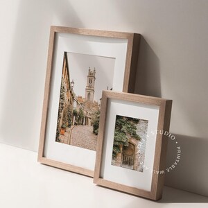 May include: Two framed photographs with wooden frames. The larger photo shows a narrow street with a church in the background. The smaller photo shows a doorway with vines growing over it.