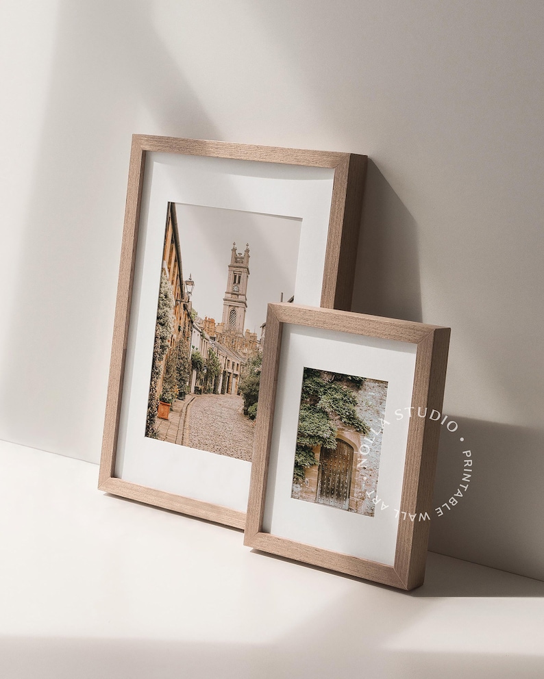 May include: Two framed photographs with wooden frames. The larger photo shows a narrow street with a church in the background. The smaller photo shows a doorway with vines growing over it.