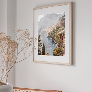 May include: A framed photograph of a coastal town nestled in a mountainside. The photo features a blue ocean, green hills, and white buildings. The frame is light wood.