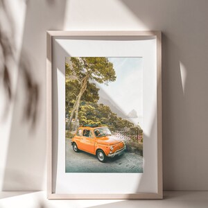 May include: A framed photograph of an orange vintage car driving on a winding road through a lush green forest. The car is parked on the side of the road with a view of the ocean in the distance.