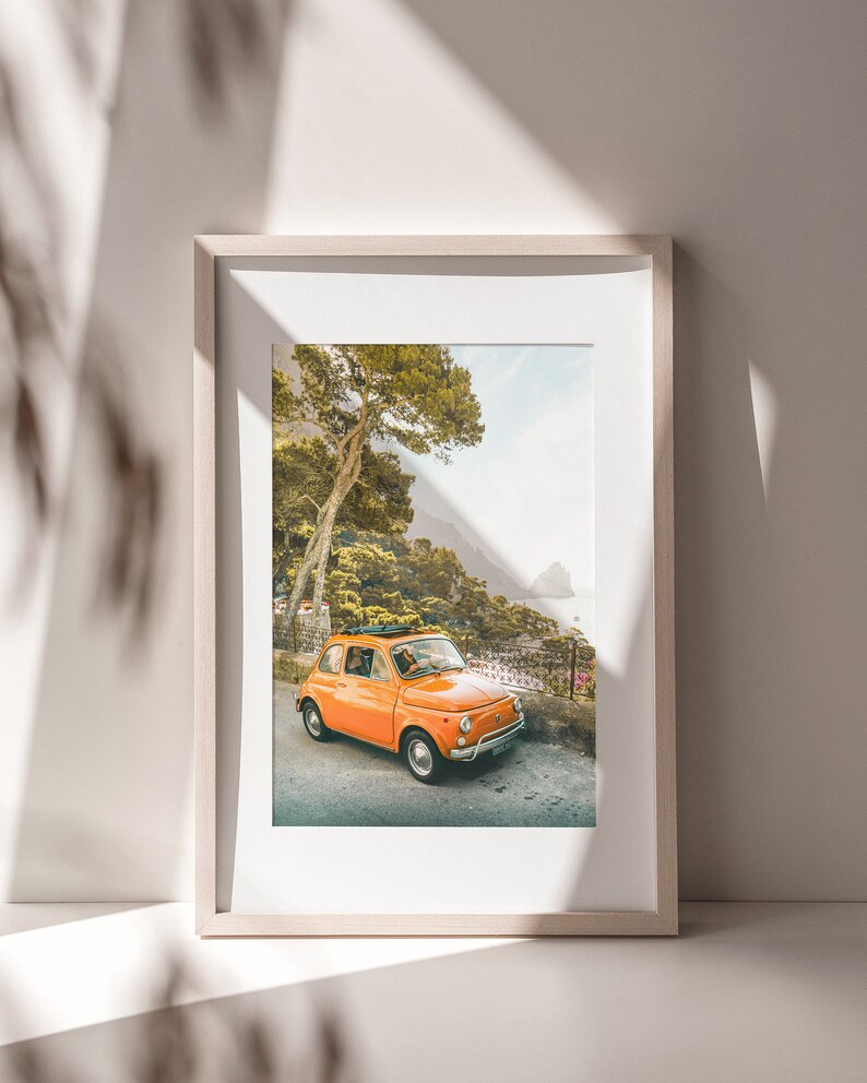 May include: A framed photograph of an orange vintage car driving on a winding road through a lush green forest. The car is parked on the side of the road with a view of the ocean in the distance.