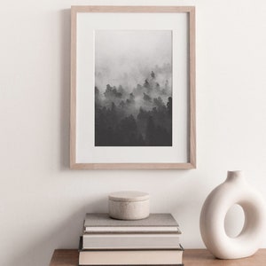 May include: A black and white photograph of a foggy forest scene in a light wood frame. The photo is displayed on a white wall above a stack of books and a white vase.