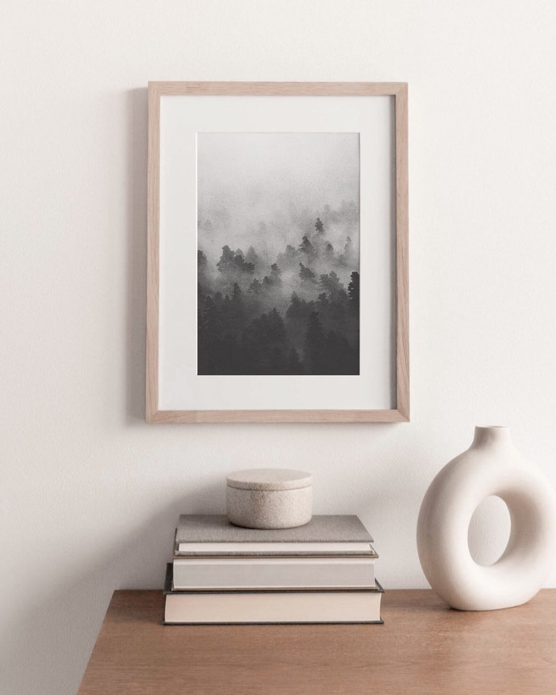 May include: A black and white photograph of a foggy forest scene in a light wood frame. The photo is displayed on a white wall above a stack of books and a white vase.