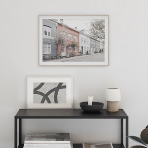 May include: A framed photograph of a row of colorful houses with a tree in the foreground. The houses are painted in shades of gray, pink, and white. The photo is hanging on a white wall above a black shelf with a candle, a bowl, and a lamp.