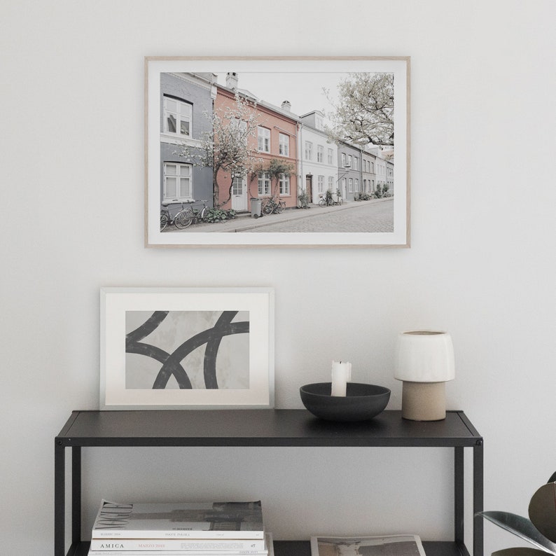 May include: A framed photograph of a row of colorful houses with a tree in the foreground. The houses are painted in shades of gray, pink, and white. The photo is hanging on a white wall above a black shelf with a candle, a bowl, and a lamp.