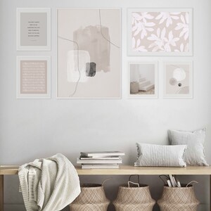 May include: A gallery wall featuring five framed prints in a minimalist style. The prints depict abstract shapes, botanical patterns, and a quote about letting go. The wall is painted a light grey and the frames are white. A wooden bench with a white throw blanket and three woven baskets sits in front of the wall.