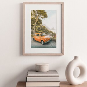 May include: A framed photograph of an orange vintage car parked on a winding road with a view of the ocean and a lush green forest in the background.