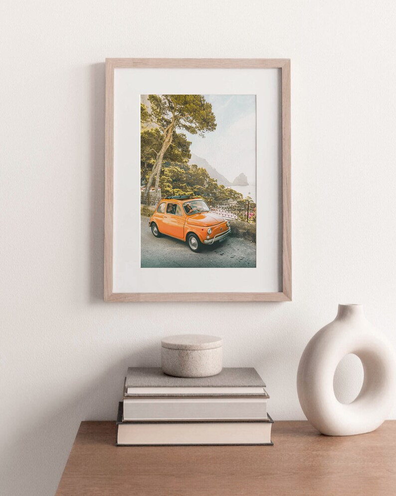 May include: A framed photograph of an orange vintage car parked on a winding road with a view of the ocean and a lush green forest in the background.