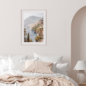 May include: A framed photograph of a coastal town nestled into a hillside overlooking the ocean. The photo is in a white frame and is hanging on a white wall above a bed with white bedding and a beige throw blanket.