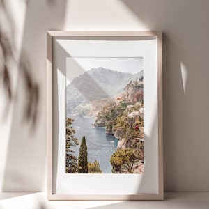 May include: A framed photograph of a coastal landscape with a view of a town nestled in the mountains overlooking a blue body of water. The photo is in a light wood frame.