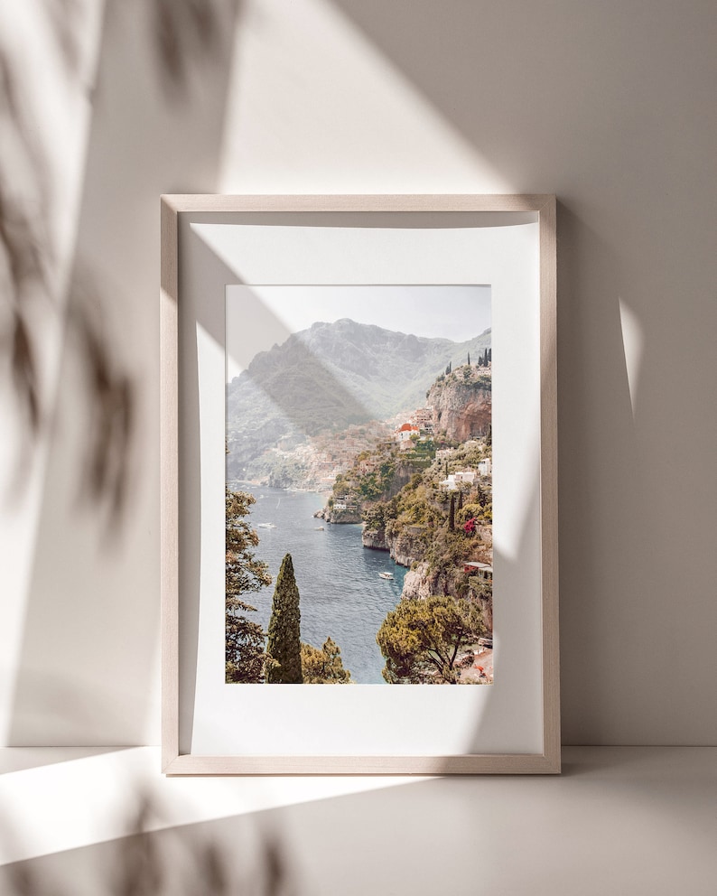 May include: A framed photograph of a coastal landscape with a view of a town nestled in the mountains overlooking a blue body of water. The photo is in a light wood frame.
