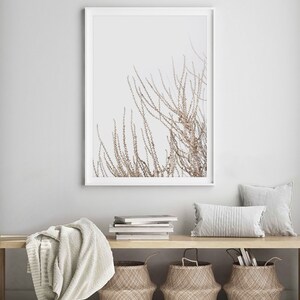 May include: A white framed print of a minimalist botanical illustration of a branch with delicate brown leaves against a white background. The print is hanging on a white wall above a wooden bench with a white throw blanket, a stack of books, and three woven baskets.