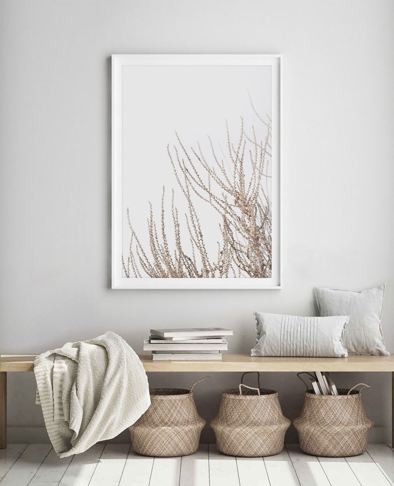 May include: A white framed print of a minimalist botanical illustration of a branch with delicate brown leaves against a white background. The print is hanging on a white wall above a wooden bench with a white throw blanket, a stack of books, and three woven baskets.