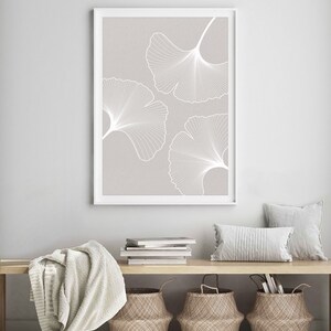 Puede incluir: Una impresi&oacute;n art&iacute;stica enmarcada con tres contornos de hojas de ginkgo blancas sobre un fondo gris claro. La obra de arte se exhibe sobre un banco de madera con cestas tejidas, cojines y una manta doblada, creando una est&eacute;tica minimalista inspirada en la naturaleza.