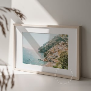 May include: A framed photograph of a coastal town built into a hillside, with a blue ocean and boats in the foreground. The image is in a light wood frame.