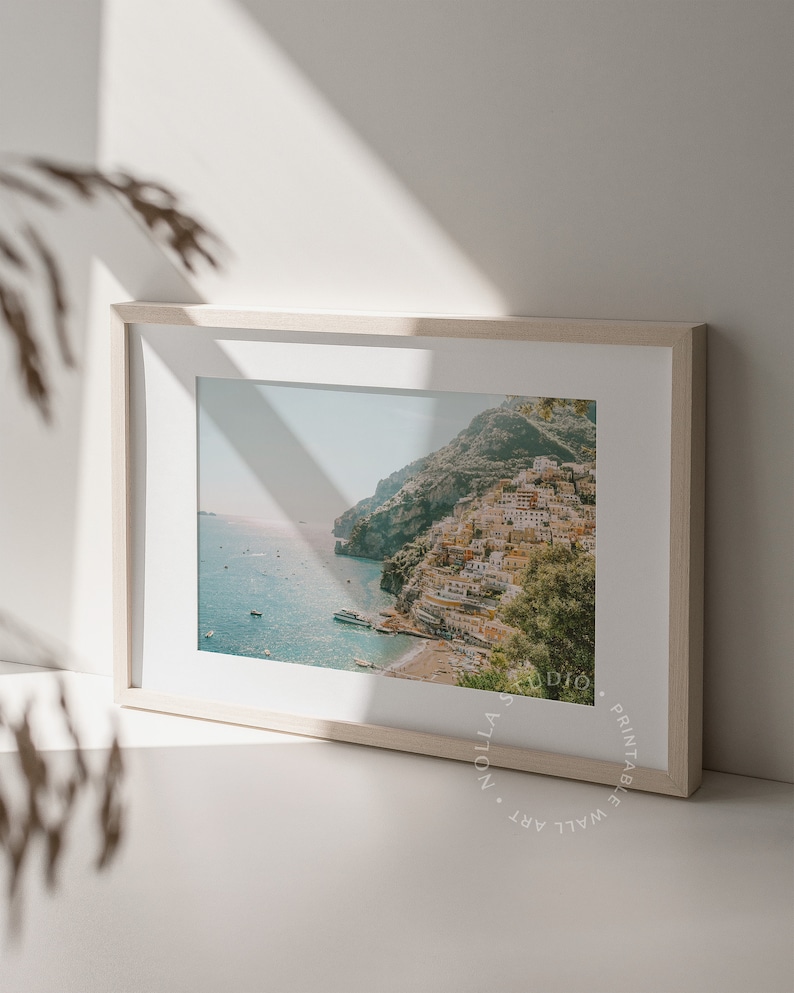 May include: A framed photograph of a coastal town built into a hillside, with a blue ocean and boats in the foreground. The image is in a light wood frame.