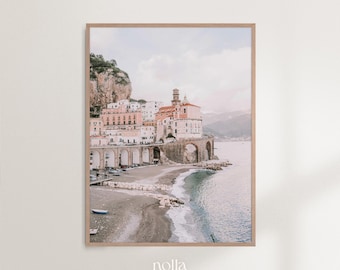 May include: A photograph of a coastal town in Italy with colorful buildings and a stone archway leading to a beach. The water is a light blue and the sky is a soft pink.