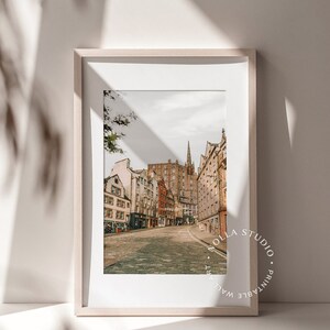 May include: A framed print of a street scene in Edinburgh, Scotland. The photo shows a cobblestone street lined with historic buildings. The print is in a light wood frame and has a white mat.