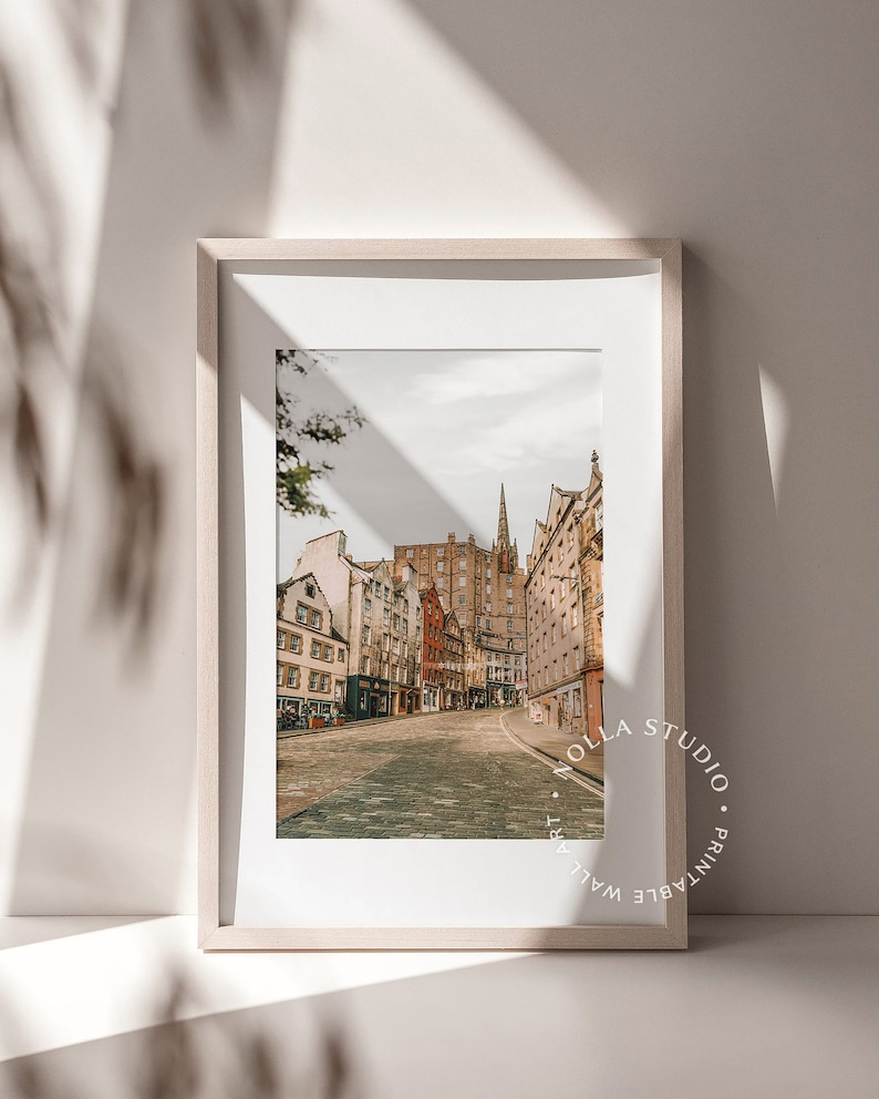 May include: A framed print of a street scene in Edinburgh, Scotland. The photo shows a cobblestone street lined with historic buildings. The print is in a light wood frame and has a white mat.