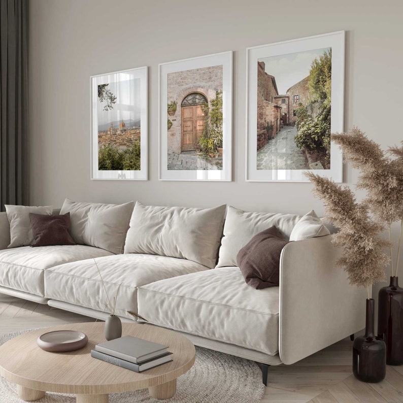 K&ouml;nnte beinhalten: Drei gerahmte Fotografien italienischer Architektur und Landschaften. Die Fotos h&auml;ngen an einer hellbeigen Wand &uuml;ber einem wei&szlig;en Sofa mit braunen Kissen.