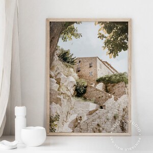 May include: A framed photograph of a stone building with a staircase leading up to it. The building is surrounded by trees and greenery. The photo is in a light brown frame.