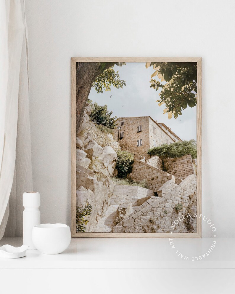 May include: A framed photograph of a stone building with a staircase leading up to it. The building is surrounded by trees and greenery. The photo is in a light brown frame.