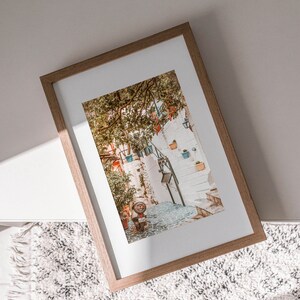 May include: A framed photograph of a narrow, cobblestone street in a European city. The street is lined with white buildings and lush greenery. A small, red scooter is parked on the street.