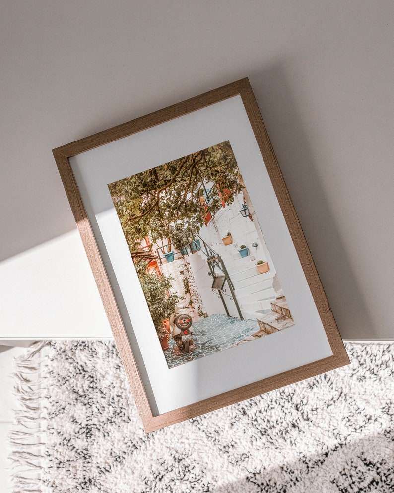 May include: A framed photograph of a narrow, cobblestone street in a European city. The street is lined with white buildings and lush greenery. A small, red scooter is parked on the street.