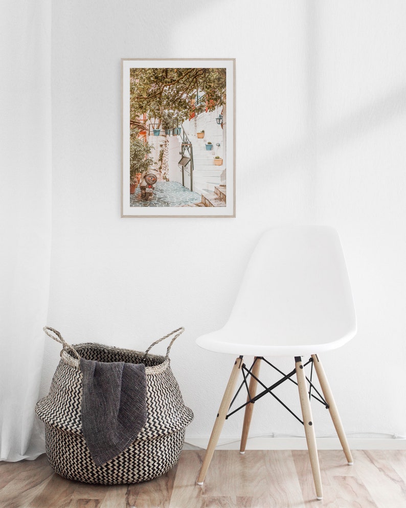 May include: A framed photograph of a white building with a stone pathway leading up to a set of stairs. The building is covered in greenery and has several potted plants hanging from the walls. The photograph is hanging on a white wall next to a white chair and a woven basket.