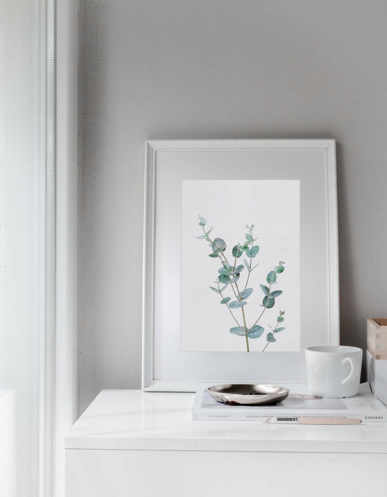 May include: A white framed print of a watercolor painting of a eucalyptus branch with green leaves against a white background. The print is hanging on a white wall above a white dresser with a white mug, a silver tray, and a magazine on top.