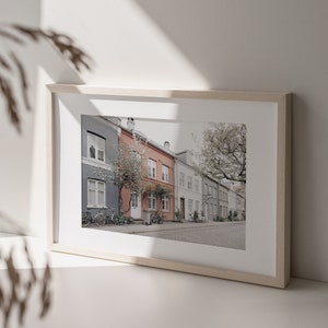 May include: A framed photograph of a row of colorful houses with bicycles parked in front of them. The houses are painted in shades of gray, pink, and white. The photo is taken on a sunny day and the street is lined with trees.