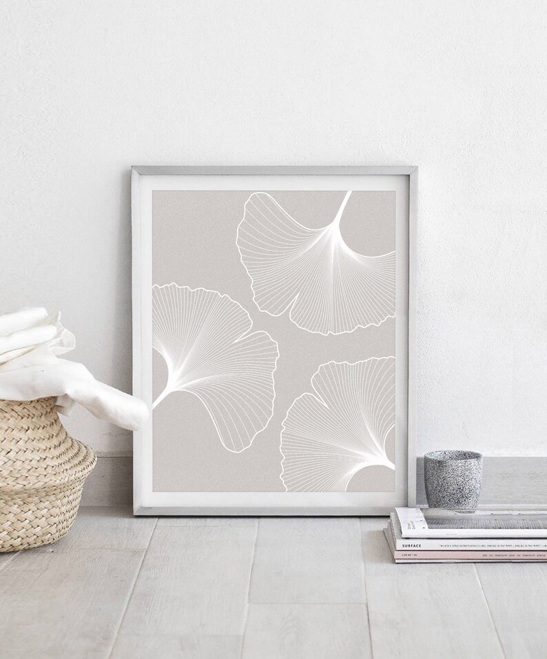 Puede incluir: Una impresi&oacute;n enmarcada con tres dise&ntilde;os de hojas de ginkgo blancas sobre un fondo taupe claro. La obra de arte est&aacute; en un marco plateado y se apoya contra una pared blanca. Se ve una cesta tejida y una peque&ntilde;a taza gris.