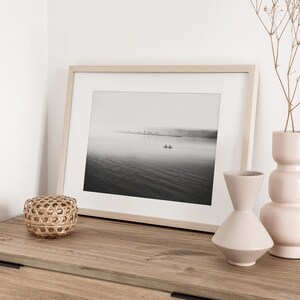 May include: A black and white photograph of a lake with a small boat in the distance. The photo is framed in a light wood frame and is displayed on a wooden shelf with two vases and a woven basket.