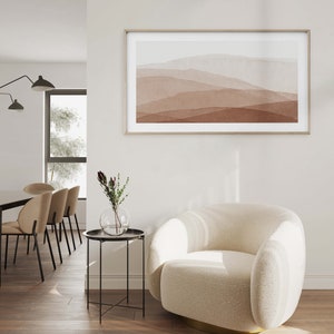 May include: A white, fluffy armchair with a gold base sits in a living room with a side table and a framed abstract painting of brown and white mountains on the wall.