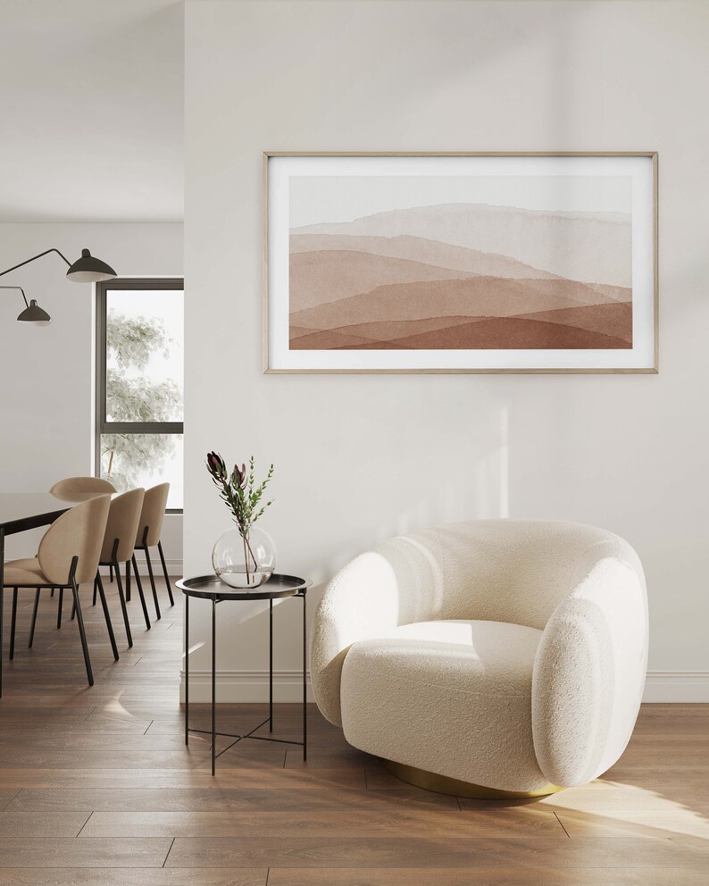 May include: A white, fluffy armchair with a gold base sits in a living room with a side table and a framed abstract painting of brown and white mountains on the wall.