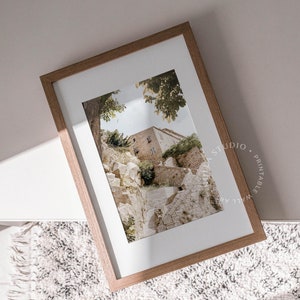 May include: A framed photograph of a stone building with a staircase leading up to it. The building is surrounded by trees and greenery. The photograph is in a light brown frame.