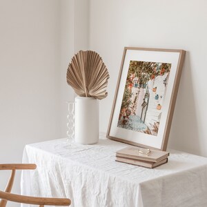 May include: A white table with a white linen tablecloth, a framed photograph of a narrow street with white buildings and steps, a dried palm leaf in a white vase, and two books on top of each other.