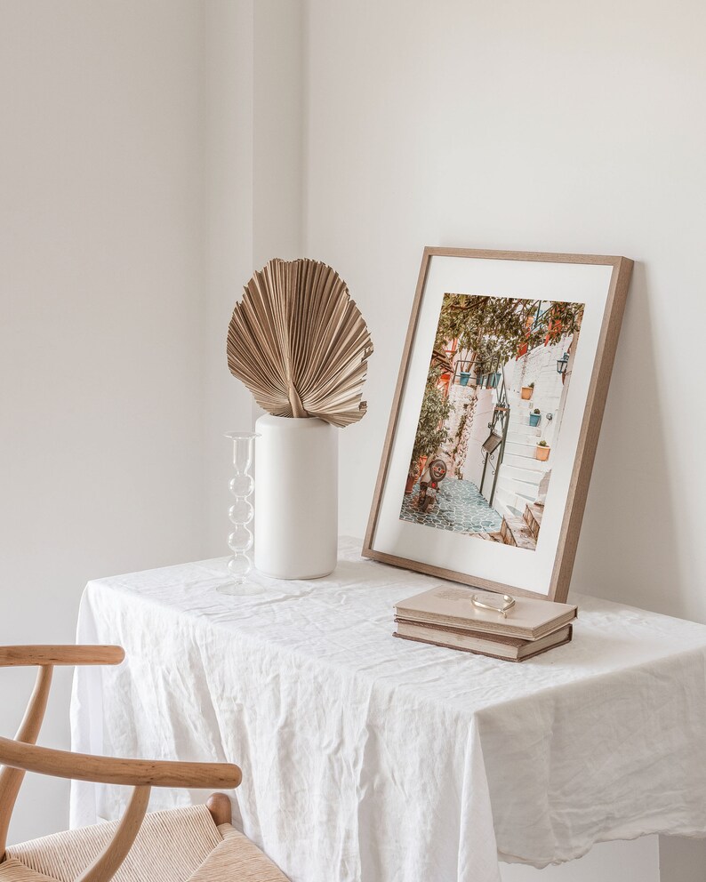 May include: A white table with a white linen tablecloth, a framed photograph of a narrow street with white buildings and steps, a dried palm leaf in a white vase, and two books on top of each other.