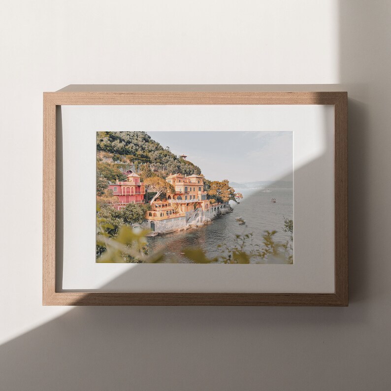 Pu&ograve; includere: Una fotografia incorniciata di un edificio colorato su una scogliera che si affaccia su un specchio d'acqua. L'edificio &egrave; un mix di giallo, arancione e rosso, ed &egrave; circondato da alberi verdi lussureggianti. L'acqua &egrave; di un blu profondo, e si vedono alcune barche in lontananza.