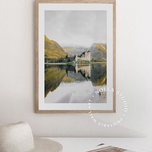 May include: A framed photograph of a castle reflected in a still lake. The castle is a gray stone structure with a pointed roof. The lake is surrounded by green hills. The sky is a light gray color. The image is framed in a light wood frame.