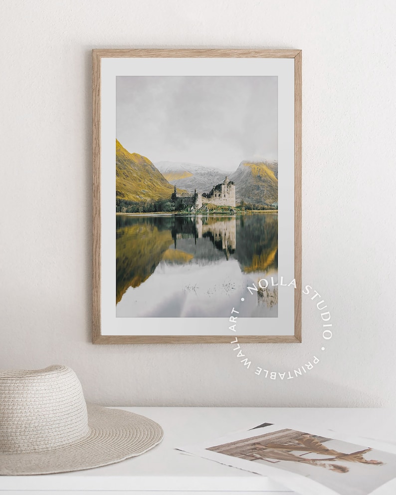 May include: A framed photograph of a castle reflected in a still lake. The castle is a gray stone structure with a pointed roof. The lake is surrounded by green hills. The sky is a light gray color. The image is framed in a light wood frame.