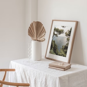 K&ouml;nnte beinhalten: Ein wei&szlig;er Tisch mit einer wei&szlig;en Tischdecke aus Leinen, ein Holzstuhl, eine Vase mit einem getrockneten Palmblatt, ein gerahmtes Foto einer Meereslandschaft und zwei &uuml;bereinander gestapelte B&uuml;cher.